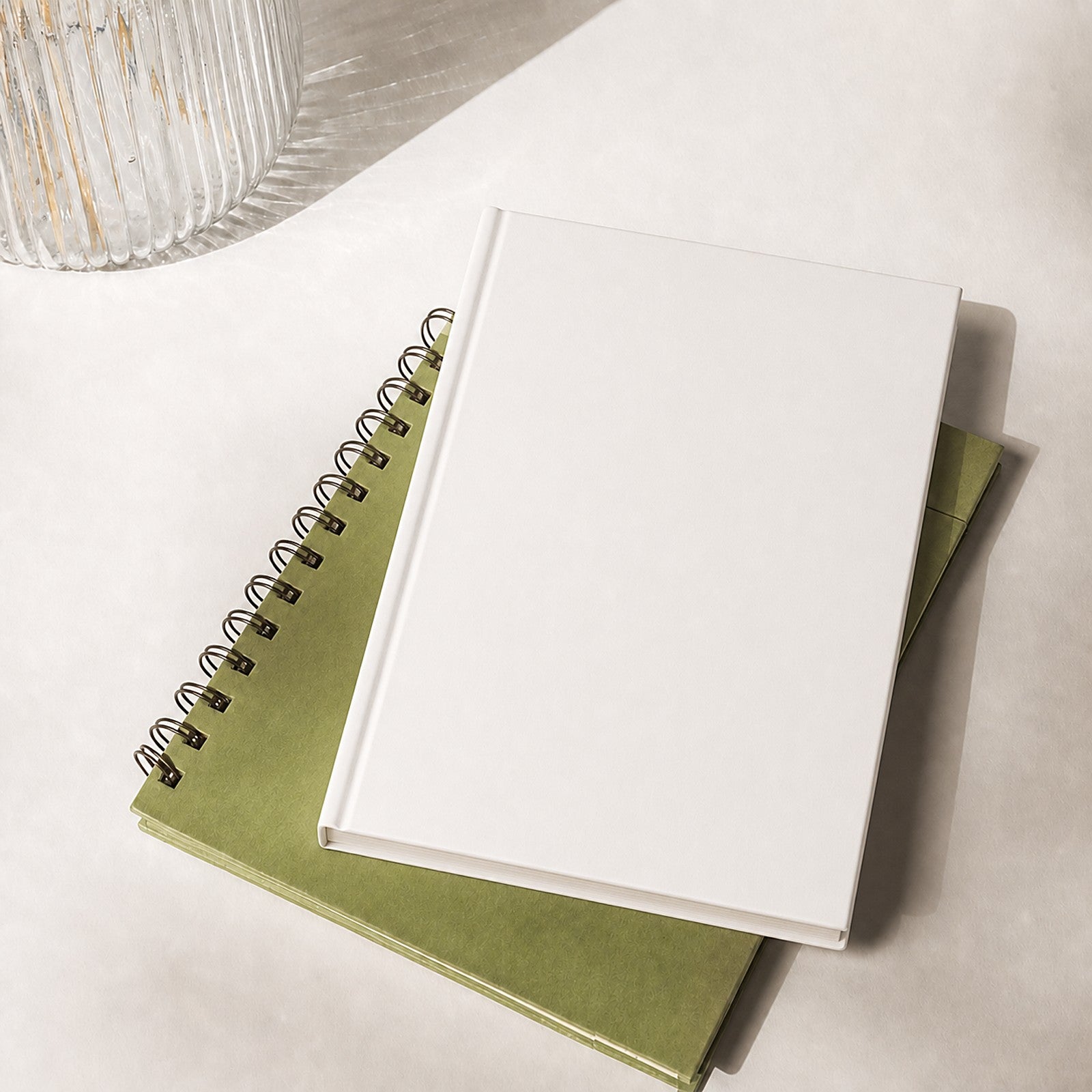 White hardcover journal lying flat on a stack of notebooks beside a ribbed glass vase